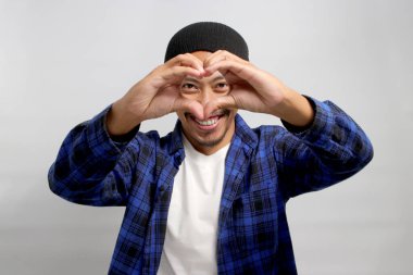 Çekici Asyalı bir adam, bereli bir şapka ve gündelik bir gömlek giyer. Elleriyle romantik bir kalp hareketi yaparken mutluluk yayar. Hassas duygularını ifade eder, beyaz arka planda durur.