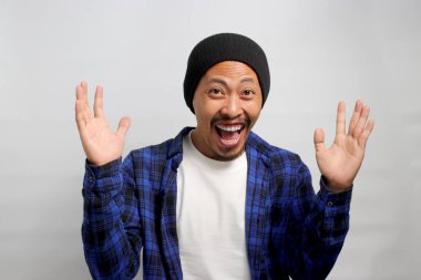 Asyalı heyecanlı genç bir adam, bereli bir şapka ve günlük bir gömlek giyerek, beyaz bir arka planda dururken sevinç ve zaferi bir yumruk pompasıyla ya da EVET hareketiyle kutluyor.