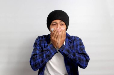 Genç Asyalı adam hapşırıyor ve burnunu bir peçeteye sümürüyor. Soğuk algınlığı, ateş ve beyaz bir zemin üzerinde izole edilmiş grip belirtileri gösteriyor. Sağlık hizmetleri ve tıbbi kavramların gösterilmesi