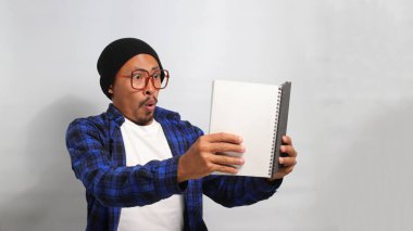 Şapka ve günlük giysiler giymiş genç bir Asyalı öğrenci, heyecan verici bir kitap okurken şaşkın ve büyülenmiş bir şekilde ağzı açık, inançsızlık içinde gösteriliyor.