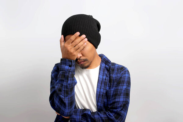 Annoyed young Asian man, dressed in beanie hat and casual shirt, is facepalming or hiding his face in frustration, feeling embarrassed due to inappropriate behavior, standing against white background