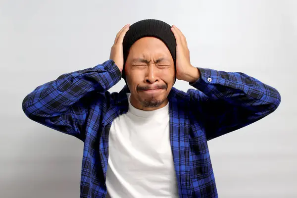 Stressed young Asian man dressed in a beanie hat and casual shirt is holding his head, possibly indicating feelings of overwhelm or distress , isolated on white background.