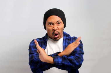 Öfkeli Asyalı genç bir adam, bereli bir şapka ve gündelik bir gömlek giyer, beyaz arka planda dururken kollarını kameraya doğru şiddetli bir şekilde çaprazlayarak reddeder ve durur.