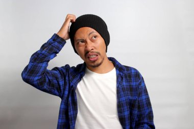 Kafası karışmış genç Asyalı bir adam, bereli bir şapka ve günlük giysiler içinde, bir çözüm ararken kafasını kaşıyor, şüphe ve kafa karışıklığını tasvir ediyor, beyaz bir arka plana karşı duruyor.