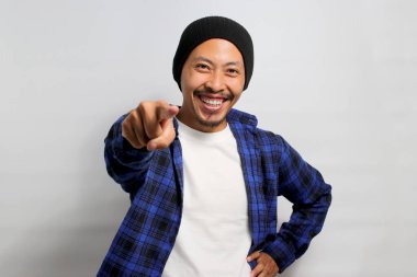 Sensin. Şapka ve günlük gömlek giymiş genç Asyalı adam kamerayı işaret ederken gülümser, insanları seçer, davet eder, adam toplar, beyaz arka planda durur.