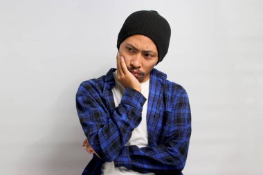 Sıkılmış genç Asyalı bir adam, bere şapkalı ve günlük kıyafetli, elini yanağında dinlendiriyor, ne yapacağını düşünüyor, sıkılmış, yorgun ve muhtemelen biraz da depresif. Beyaz arkaplanda izole.