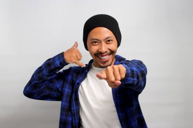 Şapka ve günlük gömlek giyen tutkulu Asyalı genç adam, enerjik bir şekilde beyaz arka planda dikilirken kameraya doğru bakarken bana bir arama hareketi yapıyor..