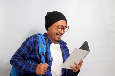 Şapka ve günlük giysiler giymiş, gözlüklü ve sırt çantası taşıyan mutlu bir Asyalı öğrenci elinde kitap tutarken beyaz bir arkaplanda duruyor.