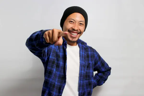 Its you. Young Asian man, dressed in a beanie hat and casual shirt, smiles while pointing his finger at the camera, choosing, inviting people, recruiting, standing over white background