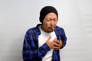 Şapkalı ve sıradan gömlekli genç Asyalı adam göğsünü sıkıca tutuyor. Muhtemel kalp krizi belirtileri gösteriyor. Şiddetli kalp ağrısı, kalp krizi ya da ağrılı kramplar.