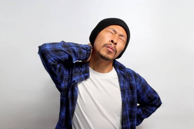 Bereli şapka ve günlük kıyafet giyen sağlıksız genç Asyalı adam boynuna masaj yapıyor. Ağrı ve uyuşukluğu ifade ediyor. Muhtemelen aşırı çalışan kaslardan kaynaklanıyor.