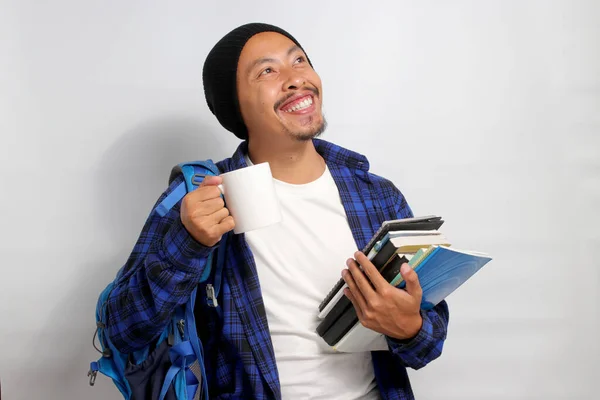 A young Asian student, dressed in casual clothes and a beanie hat, carrying a backpack and morning coffee, is prepared to study while holding books, standing against white background.