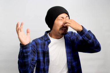 Şapka ve günlük gömlek giyen genç Asyalı adam, muhtemelen beyaz arka planda dururken kötü kokudan dolayı parmaklarıyla burnunu sıkıyor..