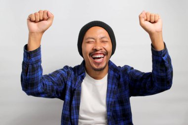 Asyalı heyecanlı genç bir adam, bereli bir şapka ve günlük bir gömlek giyerek, beyaz bir arka planda dururken sevinç ve zaferi bir yumruk pompasıyla ya da EVET hareketiyle kutluyor.