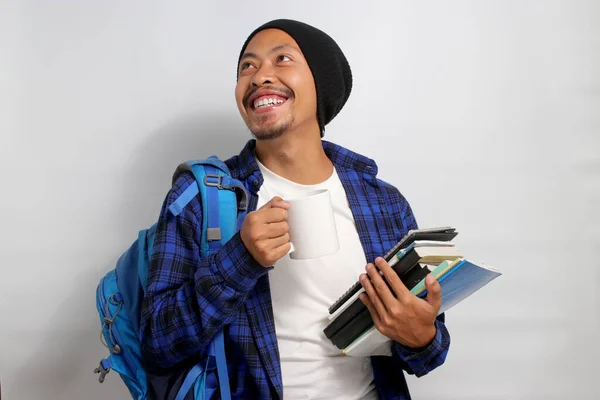 A young Asian student, dressed in casual clothes and a beanie hat, carrying a backpack and morning coffee, is prepared to study while holding books, standing against white background.