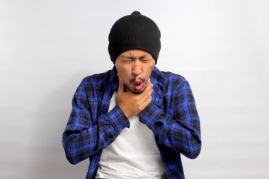 Bereli şapka ve günlük gömlek giyen genç Asyalı adam boğazında ağrı ve kuru öksürük var gibi görünüyor. Muhtemelen bir virüs veya enfeksiyon yüzünden, beyaz bir arka planda izole edilmiş..