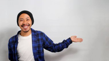 Asyalı heyecanlı genç bir adam, bereli bir şapka ve gündelik bir gömlek giyer, kollarını boş bir alana doğru uzatır, beyaz bir arka plana karşı durarak birisini kendisine katılması için davet eder.