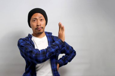 Ciddi bir Asyalı genç adam, bereli bir şapka ve günlük giysiler içinde, eli ile sıkı bir DUR hareketi yapıyor, beyaz arka plana karşı durarak reddetme, yasaklama ve uyarı işareti yapıyor.