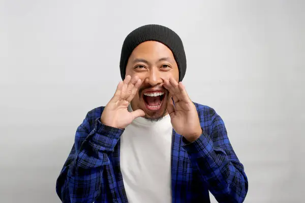 Asian man, dressed in a beanie hat and casual shirt, brings his hand near his mouth as if sharing a secret, whispering and engaging in a mischievous conversation while standing against white backdrop