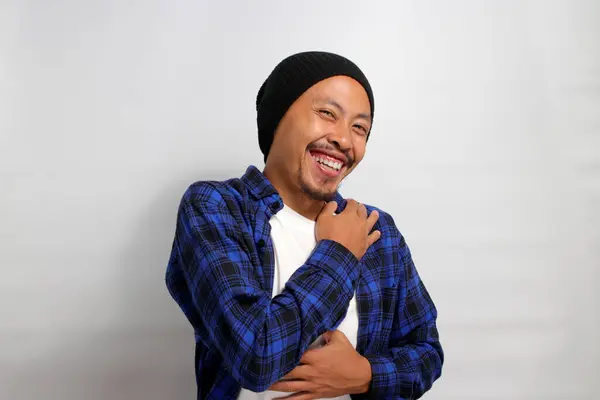 Grateful young Asian man, dressed in a beanie hat and casual clothes, expresses gratitude by placing his hand on his chest, conveying a sense of relief, happiness, and heartfelt thankfulness