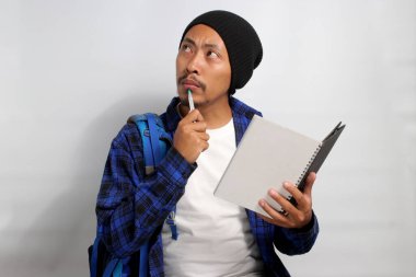 Şapka ve gündelik gömlek giymiş, sırt çantası taşıyan ve elinde defter tutan düşünceli genç bir Asyalı öğrenci, derin düşüncelere dalmış, fikirlere dalmış,