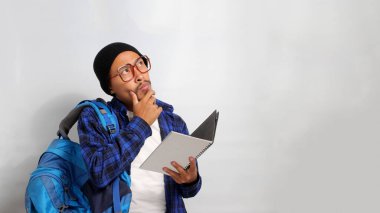 Düşünceli Asyalı bir öğrenci, gözlük takıyor, bere takıyor, gündelik giysiler giyiyor, sırt çantası taşıyor, beyaz bir arka plana yaslanıyor, bir kitap tutuyor ve elini düşünceli bir şekilde çenesine koyuyor.