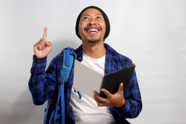 Heyecanlı Asyalı genç bir öğrenci, bereli bir şapka ve günlük bir gömlek giyiyor ve bir sırt çantası taşıyor, boş fotokopi alanını işaret ediyor, elinde bir defter, beyaz bir arkaplan üzerinde duruyor.