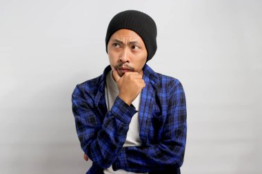 Şapka ve günlük gömlek giyen düşünceli genç Asyalı adam, elini çenesinin üzerine koyup, beyaz arka planda dururken gelecek hakkında düşünürken endişeli görünüyor..