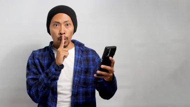 Şapka ve günlük giysiler giymiş genç bir Asyalı, parmaklarını dudaklarına dayayarak sessizlik için işaret ediyor, telefon tutarken bir sır saklamasını işaret ediyor, beyaz arka plan.