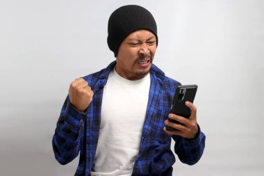 Kızgın Asyalı bir adam, bereli bir şapka ve sıradan bir gömlek giyer, kötü haberlere görünür bir şekilde tepki verir, cep telefonuna bakarken, beyaz arka planda durur.