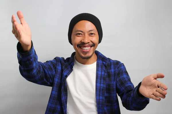 Friendly, smiling young Asian man extends his arms for a hug towards the camera, gesturing a COME HERE sign, inviting an embrace with a toothy smile while standing against white background