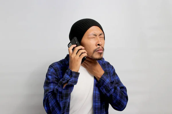 Young Asian man in a beanie hat and casual shirt is holding a mobile phone, consulting online with a doctor due to a sore throat. He is experiencing painful swallowing and discomfort