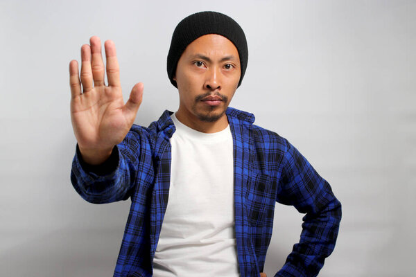 Серьезный молодой азиат, одетый в шапочку и повседневную одежду, делает жесткий жест СТОП рукой, сигнализируя об отказе, запрете и предупреждении, стоя на белом фоне