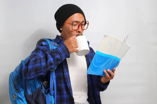 Young Asian student, dressed in casual clothes and a beanie hat, stands against a white background, holding a morning coffee while engrossed in reading a book
