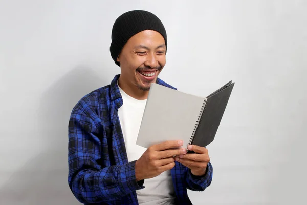 A joyful young Asian student, dressed in a beanie hat and casual shirt, is thoroughly enjoying a book, expressing happiness and a bright smile while standing against a white background