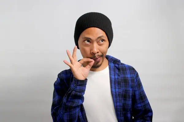 Young Asian man, dressed in a beanie hat and casual shirt, zips his lips in a promise to keep a secret, symbolically sealing his lips as if on a lock, while standing against a white background