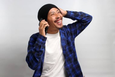 Şapka ve günlük gömlek giymiş neşeli Asyalı genç bir adam, bir telefon görüşmesi yapıyor, gülüyor ve beyaz bir arka plana karşı durarak keyifli bir sohbetin tadını çıkarıyor.