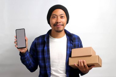 Asyalı heyecanlı genç bir adam, bereli bir şapka ve günlük kıyafetler giymiş, beyaz arka planda duran bir yığın karton kutu tutarken boş beyaz ekranlı bir akıllı telefon sergiliyor.
