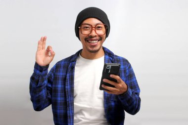 Şapka, günlük gömlek ve gözlük takan Asyalı bir adam, elinde telefon tutarken, memnuniyetini, onayını iletirken kameraya doğru 