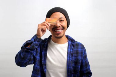 Asyalı heyecanlı genç bir adam, beresi ve günlük gömleği olan, bir gözünü kredi kartıyla kapatıyor, satın alma, tüketim, para ve ödeme kavramlarını sembolize ediyor, beyaz arka planda izole edilmiş.