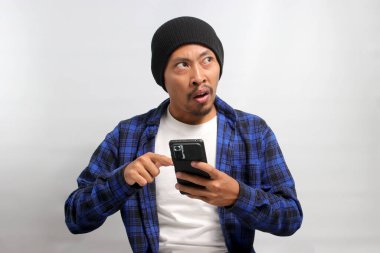 Asyalı heyecanlı genç bir adam, bereli bir şapka ve günlük bir gömlek giymiş, cep telefonunu işaret ederek beyaz bir arka plana karşı duruyor.