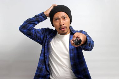 Kafası karışmış genç Asyalı bir adam, bereli bir şapka ve gündelik bir gömlek giyer, beyaz bir arka planda izole bir televizyon kumandası tutarken, şaşkınlık, şok ve şaşkınlık içinde başını kaşır.