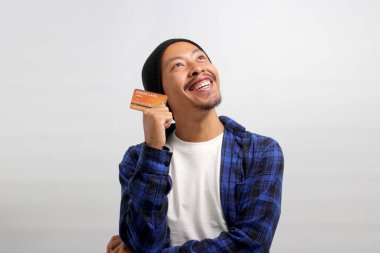 Şapka ve gündelik tişört giyen düşünceli Asyalı genç bir adam, bir kredi kartı tutuyor ve bir nebze mutluluk dolu boş bir fotokopi alanına bakıyor, kredi kartını kullanarak online alışverişler yapmayı düşünüyor.