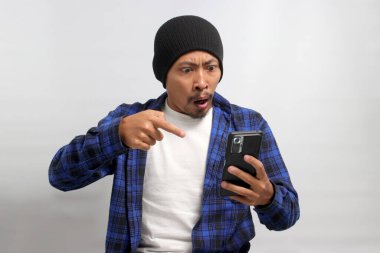 Öfkeli Asyalı bir adam, bereli bir şapka ve sıradan bir gömlek giyer, cep telefonunu işaret eder, kötü haberlere görünür bir şekilde tepki verir, yoğun öfke ve öfke gösterir, beyaz arka planda durur.