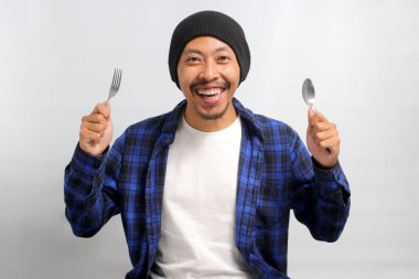 Şapkalı ve rahat gömlekli neşeli Asyalı bir adam elinde çatal ve kaşıkla kameraya bakıp heyecanla gülümserken lezzetli bir yemek bekliyor.