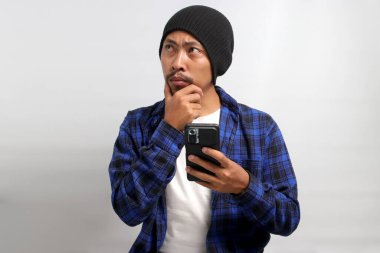 Kafası karışmış ve kafası karışmış Asyalı bir adam, bereli şapka ve günlük gömlek giymiş, cep telefonundaki haberlere tepki veriyor, kafa karışıklığı, mutsuzluk ve derin tefekkür sergiliyor, beyaz arka planda izole edilmiş.