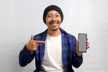 Heyecanlı Asyalı bir adam, bereli bir şapka ve günlük bir gömlek giymiş, boş beyaz ekranlı bir cep telefonuna bakıyor, yeni bir uygulama veya özel bir teklif öneriyor ya da öneriyor.