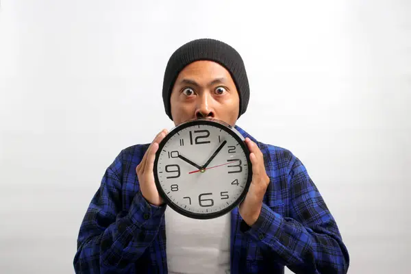 An Asian man, sporting a beanie hat and casual shirt, holds a clock in front of his face, looking at the camera with a surprised and humorous expression, while standing against a white background