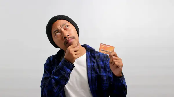 Pensive young Asian man, dressed in a beanie hat and casual shirt, holds a credit card and gazes at an empty copy space with a hint of happiness, contemplating online purchases using the credit card