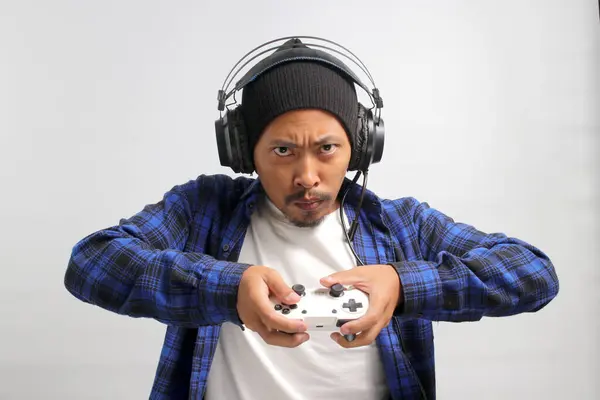 An angry and displeased young Asian man, dressed in a beanie hat and casual shirt, appears stressed while playing a video game using a gamepad on a game console, isolated on white background